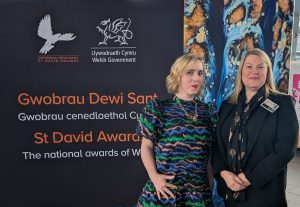 Alt Text: CEO Andrea Wayman, and ELITE Group Admin Manager Erika James, stand in front of a banner for St David Awards. Both look extremely proud. The banner reads "Gwobrau Dewi Sant, Gwobrau cenedlaethol Cymru, St David Awards, The national awards of Wales." The banner also features logos for "Llywodraeth Cymru Welsh Government" the bird in flight logo for the Saint David's Award.