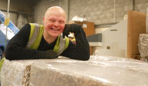 A smiling young man in high vis rests on bales of cardboard.