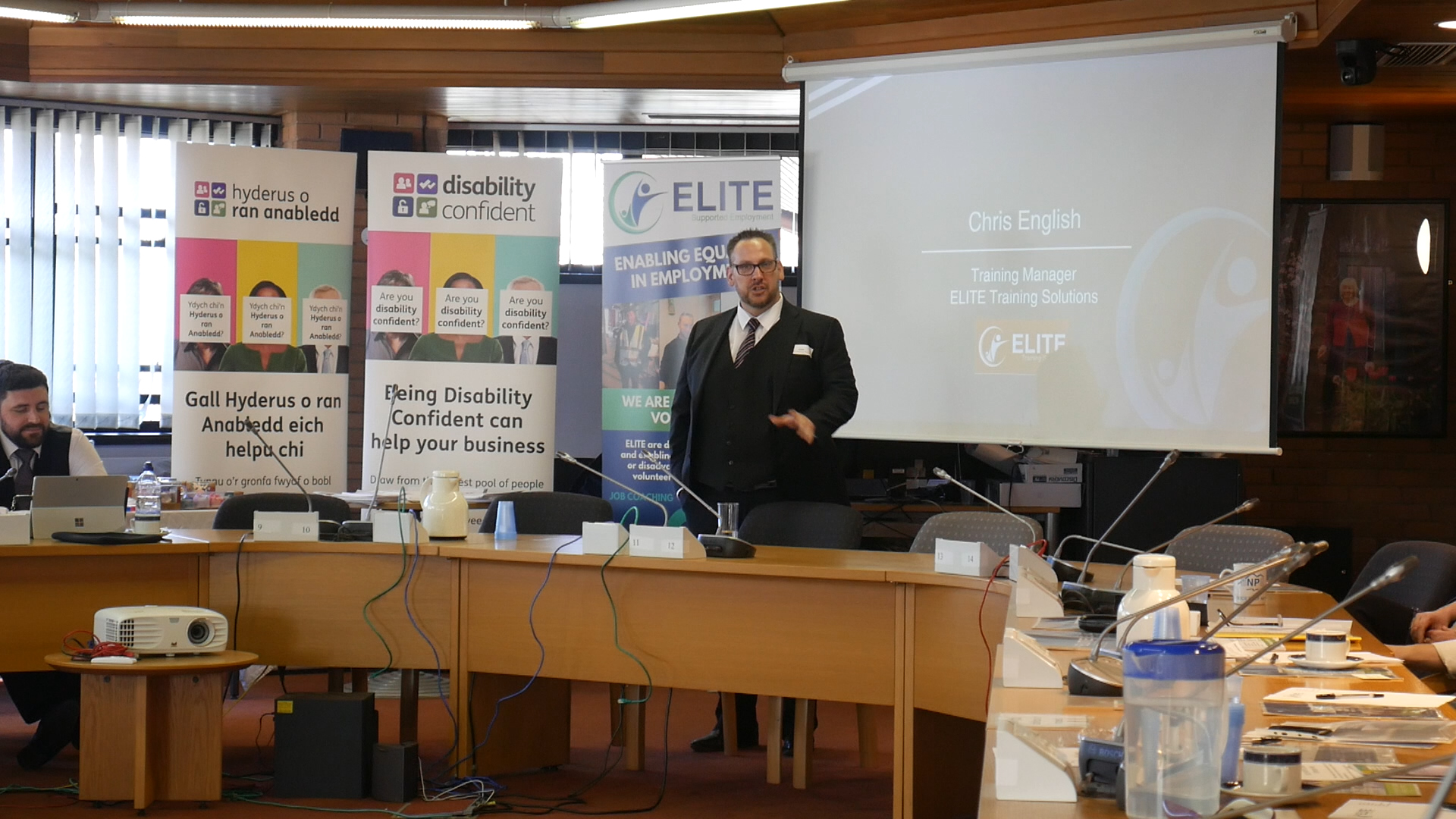 Chris, Training Manager at ELITE Training Solutions, presents in a conference room with banners displaying 'Disability Confident,' 'Hyderus o ran anabledd,' and 'ELITE.' The room is arranged in a U-shape with laptops and training materials on the tables."