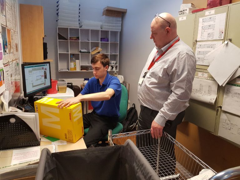 Two individuals in an office or storage room. One is seated at a desk using a computer and handling a large yellow box marked with an 'M'; the other stands nearby observing. The room contains shelves, office supplies, a shopping cart, and pinned papers.