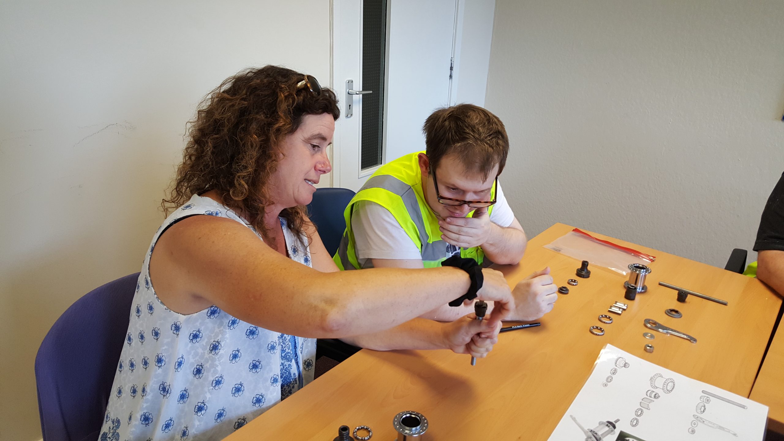 Two individuals seated at a table assembling mechanical parts. One person demonstrates tool use while the other, wearing a high-visibility vest, observes. Mechanical components and an instruction sheet with diagrams are spread out on the table