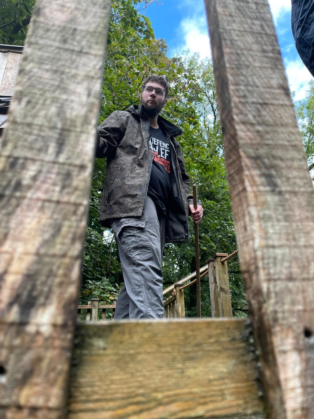 A young person is standing outside on a wooden platform between two trees. He's about to take a step somewhere,