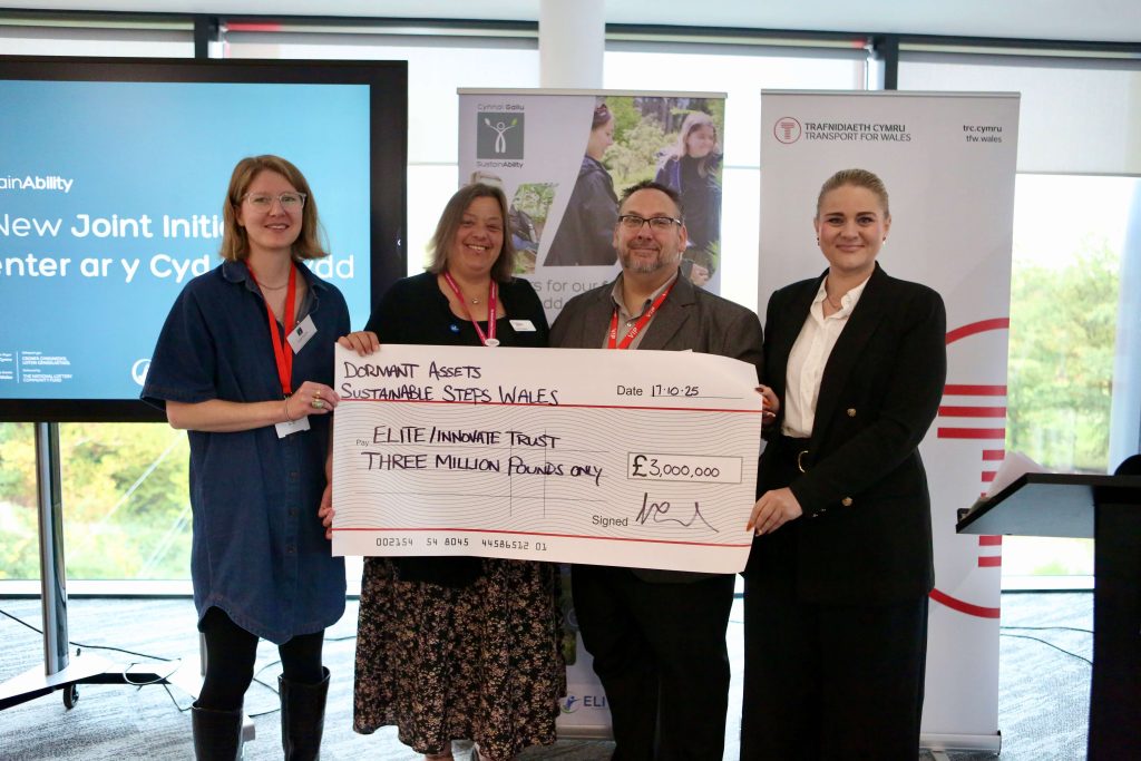 Innovate Trust, ELITE, Lottery staff members stand indoors alongside the local MP for Pontypridd, they are holding a large ceremonial check made out to 'Sustainable Steps Wales' from 'ELITE/Innovate Trust' for £3 million, dated 17-10-23. Behind them are banners and a screen displaying information about the new joint initiative."