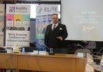 Chris, Training Manager at ELITE Training Solutions, presents in a conference room with banners displaying 'Disability Confident,' 'Hyderus o ran anabledd,' and 'ELITE.' The room is arranged in a U-shape with laptops and training materials on the tables."