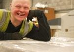 A smiling young man in high vis rests on bales of cardboard.
